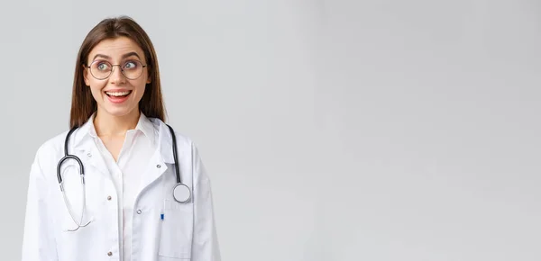 Trabajadores sanitarios, medicina, seguros y concepto de pandemia covid-19. Feliz médico optimista mujer, médico con estetoscopio en uniformes blancos y gafas, mira esquina superior izquierda con sonrisa optimista — Foto de Stock