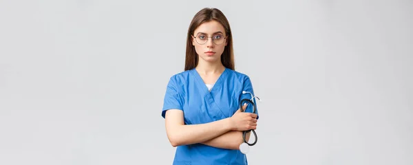 Egészségügyi dolgozók, orvostudomány, biztosítás és a kovid-19 világjárvány koncepciója. Komoly és határozott, hivatásos női ápolónő, orvos kék köpenyben, kezében sztetoszkóppal, szemüveget visel, magabiztosnak látszik — Stock Fotó