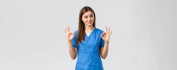 Healthcare workers, prevent virus and medicine concept. Attractive confident female nurse, doctor in blue scrubs, smirk show okay sign, guarantee quality of service in clinic, recommend medicine — Stock Photo, Image