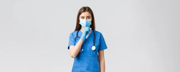 Covid-19, het voorkomen van virus, gezondheid, gezondheidswerkers en quarantaine concept. Praat wat zachter. Serieuze vrouwelijke verpleegster in blauwe scrubs, medisch masker en handschoenen, shushing, druk vinger op de lippen — Stockfoto