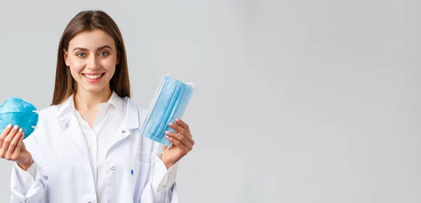 Covid-19, forebyggelse af virus, sundhedspersonale koncept. Smilende attraktiv kvindelig læge i hvide scrubs, der viser to varianter af personlige værnemidler, viser respirator og medicinsk maske - Stock-foto