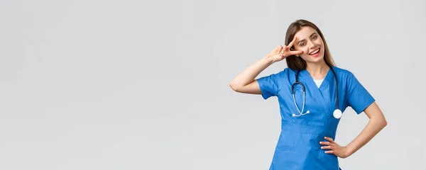 Egészségügyi dolgozók, egészségügy, kovid-19 és oltási koncepció. Vidám vonzó női nővér vagy orvos kék köpenyben, sztetoszkóp, mutatják a béke jele, maradj optimista, álló szürke háttér — Stock Fotó