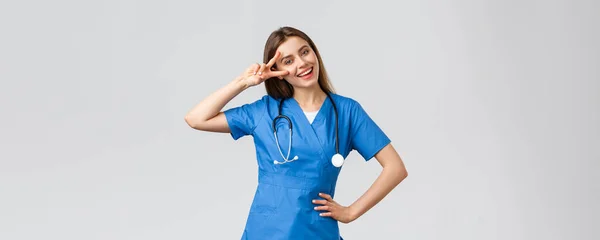 Egészségügyi dolgozók, egészségügy, kovid-19 és oltási koncepció. Vidám vonzó női nővér vagy orvos kék köpenyben, sztetoszkóp, mutatják a béke jele, maradj optimista, álló szürke háttér — Stock Fotó