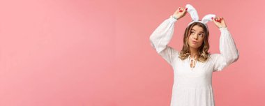 Tatiller, bahar ve parti konsepti. Tavşan kulaklı aptal sarışın kızın portresi, hayal kuruyor, gülümsüyor, beyaz elbise giyiyor, pembe arka planda kaygısız duruyor.