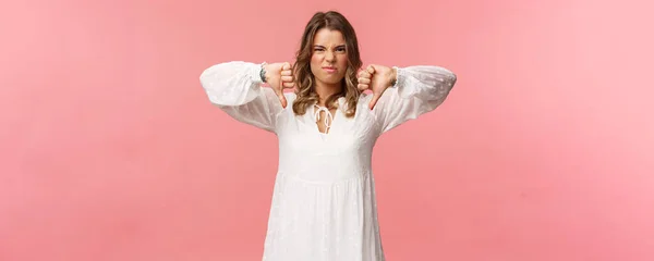Portrait of picky young blond caucasian woman in white dress over pink background expressing dislike, show thumbs-down and grimacing in aversion, upset with bad quality — Stok Foto