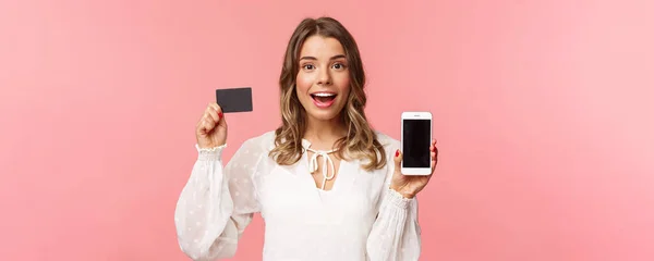 Concepto de finanzas, compras y tecnología. Retrato de cerca de la chica atractiva rubia emocionada en vestido blanco, mostrando la tarjeta de crédito y el teléfono móvil, anunciar tienda en línea, aplicación para teléfonos inteligentes — Foto de Stock