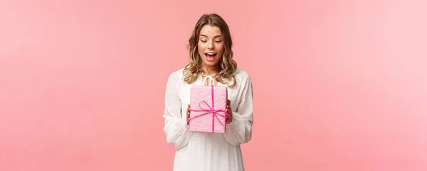 Feriados, celebração e conceito de mulheres. Retrato de menina loira encantadora surpresa receber presente surpresa, segurando presente na caixa rosa olhar para ele divertido, curioso o que está dentro, fundo do estúdio — Fotografia de Stock