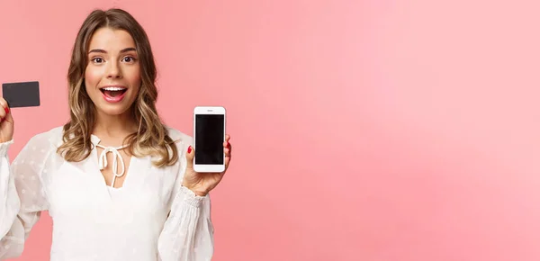 Ekonomi, shopping och teknik. Närbild porträtt av upphetsad blond attraktiv flicka i vit klänning, visar kreditkort och mobiltelefon, annonsera online-butik, smartphone app — Stockfoto