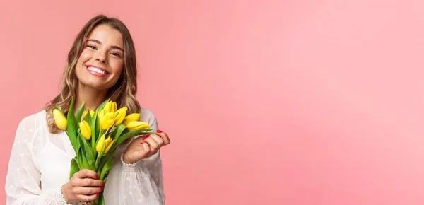 Frühling, Glück und Feiern. Nahaufnahme eines schönen, glücklichen jungen blonden Mädchens in weißem Kleid, das nach dem perfekten Date tagträumt, gelbe Tulpen in die Kamera lächelt, rosa Hintergrund — Stockfoto