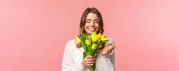 Frühling, Glück und Feiern. Nahaufnahme Porträt eines schönen, romantischen blonden Mädchens, das den Geruch schöner gelber Tulpen schnuppert, die Augen schließt und glücklich lächelt, auf rosa Hintergrund stehend — Stockfoto