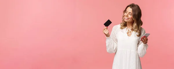 Retrato de mujer rubia joven y guapa feliz en vestido blanco, mirando satisfecho y satisfecho en la tarjeta de crédito, sosteniendo el teléfono inteligente, recomiendan comprar en línea, concepto de compras por Internet — Foto de Stock