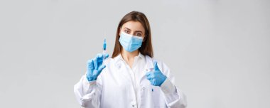 Covid-19, virüsü önleme, sağlık çalışanları ve karantina konsepti. Tıbbi maske ve lastik eldiven takmış kararlı genç doktor, önlükler baş parmak ve şırıngada koronavirüs aşısı gösteriyor.