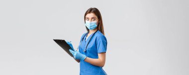 Covid-19, virüsün önlenmesi, sağlık, sağlık çalışanları ve karantina konsepti. Şok olmuş ve ürkmüş kadın hemşire ya da doktor mavi önlüklü, tıbbi maskeli, elinde pano bakışlarıyla kameraya şaşırmış.