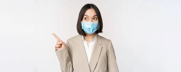 Femme d'affaires enthousiaste pointant les doigts vers la gauche, portant un masque médical de la pandémie de covide-19, debout sur fond blanc — Photo