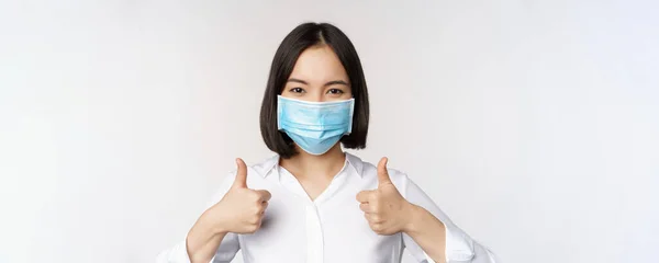 Porträtt av leende asiatiska kontor dam i medicinsk ansiktsmask, visar tummen upp, rekommendera smth, står över vit bakgrund — Stockfoto