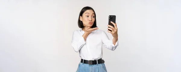 アジアの可愛い女の子のビデオチャットの画像,カメラでエアキスを送信,スマートフォン上のアプリフィルターで自撮りを取ります,白い背景の上に立って — ストック写真