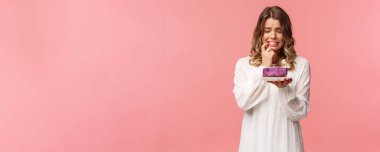 Tatiller, bahar ve parti konsepti. Tedirgin sarışın bir kızın portresi, dudak ısırması lezzetli pasta yeme arzusuna karşı koyamaz. Tereddütlü bir ifadeyle tatlıya bakar, endişeli suratını buruşturur.