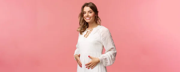 Maternidade, mulheres e conceito de beleza. Mulher loira sorridente macia e fofa em vestido branco sentindo felicidade e amor, tocando na barriga como esperando criança, estando grávida, fundo rosa — Fotografia de Stock