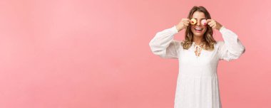 Tatiller, bahar ve parti konsepti. Beyaz elbiseli, kaygısız, komik ve tatlı sarışın bir kızın portresi. Gözlerinde iki badem kurabiyesi var ve mutlu gülümsüyor. Lezzetli tatlılar yemek gibi, pembe arka plan.