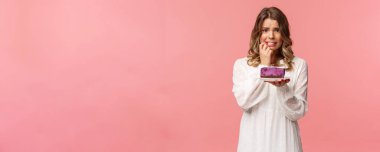 Tatiller, bahar ve parti konsepti. Telaşlı, endişeli genç sarışın kız kekini yerken ya da yemezken diyette olmak, sağlıklı kalmaya çalışmak, tatlıyı tutmak, gergin bir şekilde parmağını ısırmak, pembe arka plan.