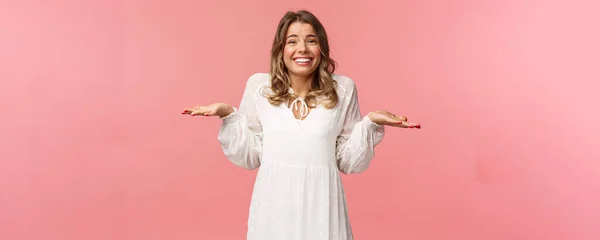 Conceito de beleza, moda e mulheres. Desculpa não saber, não posso dizer. alegre bonito bobo loiro menina 20s, usar vestido branco, encolher os ombros com as mãos lateralmente fazer estranho sorriso, desculpas, fundo rosa — Fotografia de Stock