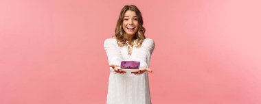 Tatiller, bahar ve parti konsepti. Şefkatli, romantik ve tatlı sarışın kız arkadaş lezzetli bir tatlı hazırladı, denemenizi istiyor, insanlara kek veriyor ve gülümsüyor, pembe arka plan