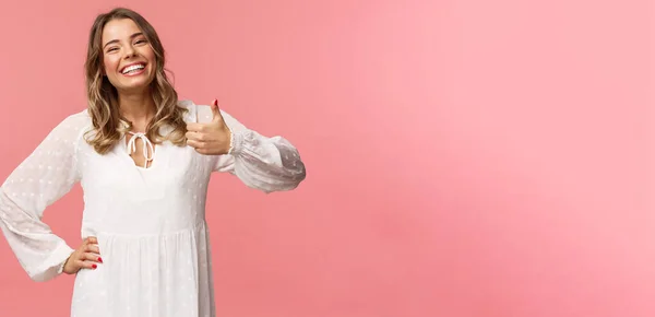 Entusiástico linda jovem mulher em vestido bonito branco, mostrar polegar-up na aprovação, recomendar o produto, sorrindo satisfeito, deixar comentário positivo, impressão, de pé fundo rosa — Fotografia de Stock