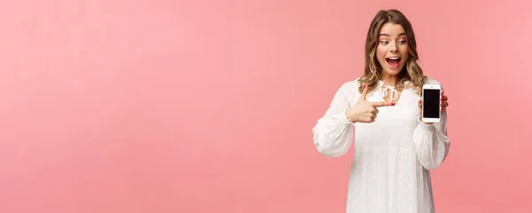 Retrato de animado e surpreso, feliz mulher loira em vestido branco, apontando o dedo para a tela do telefone móvel, olhando para o smartphone com sorriso satisfeito e surpreso, stand pink background — Fotografia de Stock