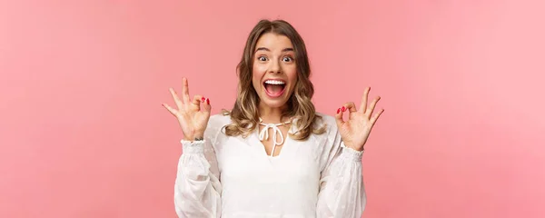 Retrato de excitado y optimista, alegre chica rubia en vestido blanco bailando y mostrando signos de bien con sonrisa radiante feliz, emocionado por el festival de música, película impresionante tasa, recomendar producto — Foto de Stock