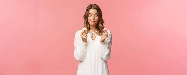 Férias, primavera e festa conceito. Retrato de linda menina loira romântica em vestido branco, lambendo os lábios como tentador para comer deliciosa sobremesa, segurando dois macarons e olhar satisfeito, fundo rosa — Fotografia de Stock