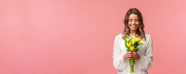 Tatiller, güzellik ve bahar konsepti. Beyaz elbiseli, sarı laleli, güzel çiçeklere bakan, gülümseyen ve kızaran pembe arka planlı güzel, romantik sarışın kızın portresi.