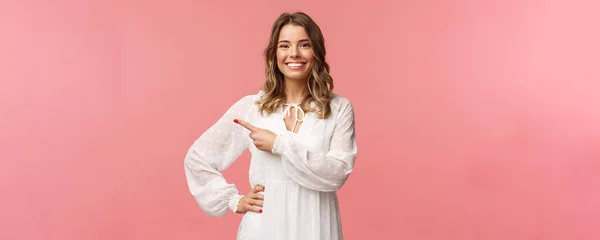 Agradável mulher loira sorridente de boa aparência com cabelo ondulado curto loiro, vestido macio branco, olhando câmera e apontando o dedo esquerdo para mostrar-lhe o melhor preço, boa oferta, conceito promocional, fundo rosa — Fotografia de Stock