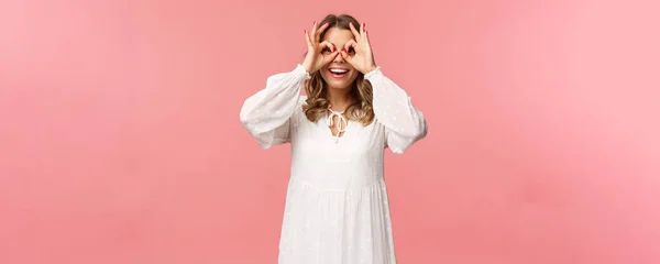 Retrato de sonho bonito e engraçado jovem loira vendo algo interessante, olhando de sinais ok como fazer máscara de vidro com os dedos sobre os olhos, sorrindo divertido, fundo rosa — Fotografia de Stock