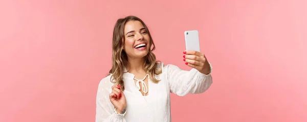 Närbild porträtt av glada optimistiska leende blond flicka, bär vit klänning, skrattar som inspelningsvideo, ringer vän på mobilen ansökan, ta foto, selfie på smartphone, rosa bakgrund — Stockfoto