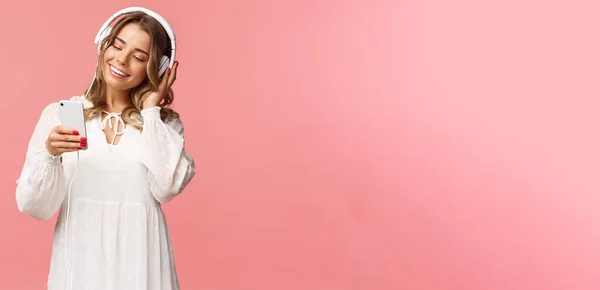 Retrato de sonho bonito e macio menina loira em vestido branco, segurando smartphone, ouvir música em fones de ouvido, sorrindo para exibir telefone celular como escolher playlist para o humor da primavera, fundo rosa — Fotografia de Stock