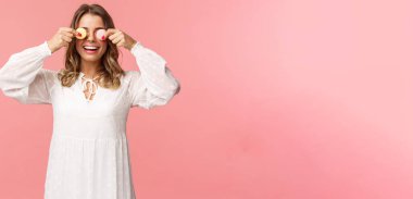 Tatiller, bahar ve parti konsepti. Beyaz elbiseli, kaygısız, komik ve tatlı sarışın bir kızın portresi. Gözlerinde iki badem kurabiyesi var ve mutlu gülümsüyor. Lezzetli tatlılar yemek gibi, pembe arka plan.