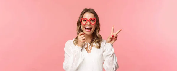 Spring, happiness and celebration concept. Close-up portrait of upbeat blond girl at party, wear white dress holding heart-shaped glasses mask over eyes and make peace sign cheerful — Stock Photo, Image