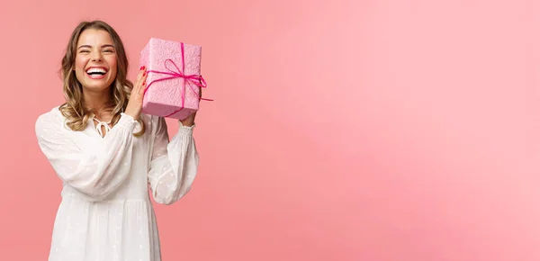 Fiestas, celebración y concepto de mujer. Retrato de la feliz chica rubia carismática sacudiendo caja de regalo preguntándose qué hay dentro como celebración de cumpleaños, recibir regalos del b-day, fondo rosa —  Fotos de Stock