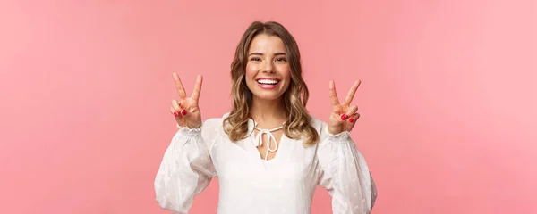 Close-up portret optymistyczne kawaii młoda blondynka z pięknym uśmiechem promieni, pokazując znaki pokoju i patrząc aparat z pozytywnym nastawieniem, korzystających wiosna, biała sukienka, różowe tło — Zdjęcie stockowe