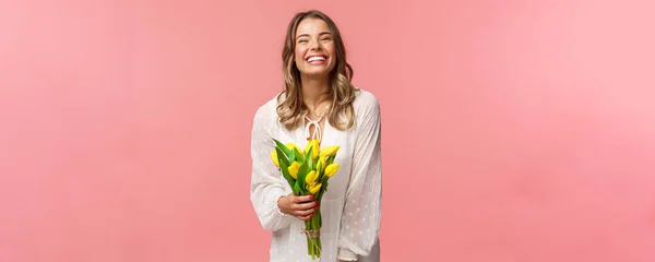 Liburan, keindahan dan konsep musim semi. riang gembira menarik gadis pirang dengan gaun putih memegang tulip kuning dan tertawa dengan senang hati ekspresi, berdiri latar belakang merah muda, memiliki tanggal romantis — Stok Foto