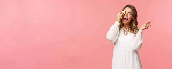 Vacaciones, primavera y concepto de fiesta. Retrato de niña despreocupada, tierna y femenina en vestido blanco, sosteniendo macarrones postre sobre el ojo, sintiéndose alegre, comiendo dulces, sabrosos postres, fondo rosa —  Fotos de Stock