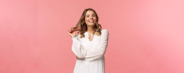 Güzel ve kendine güvenen genç, sarışın, hoş, şık, beyaz elbiseli, saçlarına dokunan ve gülümseyen, romantik randevu için giyinen, kadınlar ve güzellik konsepti, pembe arka plan