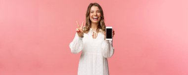 Kawai 'nin portresi iyimser ve mutlu beyaz elbiseli genç kız, cep telefonu ekranını ve barış işaretini göster, gülen ve mutlu hisseden harika bir uygulama, yararlı bir bağlantı, pembe arka plan