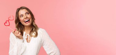 Bahar, mutluluk ve kutlama konsepti. Güzel sarışın kızın yakın plan portresi, kalp şeklinde bir maske takıyor ve hayal kuruyor, mutlu bir gülümsemeyle düşünceli bir şekilde yukarı bakıyor.