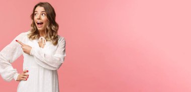 Heyecanlı ve şaşkın sarışın beyaz kız, kawaii elbisesi içinde, ağzı açık huşu içinde, heyecan ve şaşkınlıkla işaret ederek sol tarafa bakıyor, harika bir güzellik ürünü, pembe arka plan