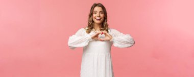 Beauty, fashion and women concept. Romantic passionate blond european female in white cute dress, make heart sign near chest and smiling at camera excited, show sympathy clipart
