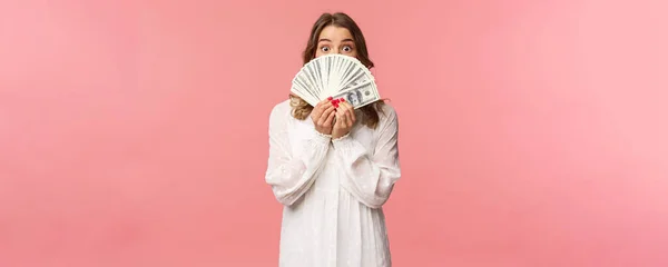 Ritratto di stupito ed eccitato carino ragazza bionda femminile in abito bianco, in possesso di dollari sul viso, guardando da sotto contanti alla fotocamera con espressione sorpresa, stand sfondo rosa — Foto Stock