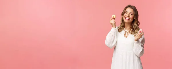Vacaciones, primavera y concepto de fiesta. Retrato de mujer rubia tierna y encantadora en vestido blanco, bailando alegremente con dos macarrones, sonriendo feliz comiendo delicioso postre, comida sabrosa, fondo rosa — Foto de Stock