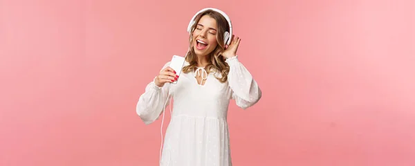 Portrait of beautiful blond caucasian female in white dress, listening music in headphones, close eyes and smiling, using mobile phone like microphone, singing along favorite song, pink background — Stock Photo, Image
