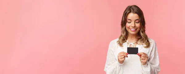Close-up retrato de menina loira animada e divertida em vestido branco, segurando cartão de crédito e sorrindo emocionado, não pode resistir à tentação de comprar algo, desperdiçar dinheiro compras on-line, fundo rosa — Fotografia de Stock